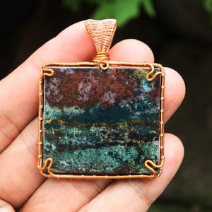 Puede incluir: Colgante cuadrado con marco y asa de alambre de cobre. El colgante presenta una piedra con una mezcla de colores verdes, marrones y rojos. El alambre está intrincadamente tejido alrededor de la piedra, creando una joya única.
