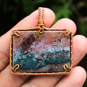 Puede incluir: Un colgante envuelto en alambre de cobre con una piedra de ágata colorida con tonos de marrón, rosa, verde y blanco. La piedra está engastada en un marco cuadrado.