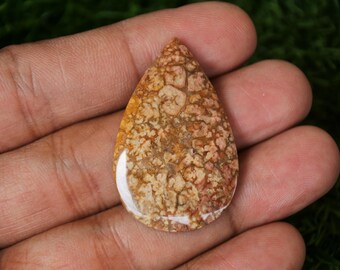 May include: A teardrop-shaped cabochon gemstone with a brown, white and orange pattern. The stone has a smooth, polished surface.