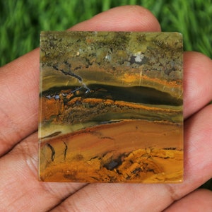 May include: A square, polished, multi-coloured gemstone with brown, yellow, green, and black banding. The gemstone has a smooth, polished surface.