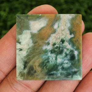 May include: Square gemstone with a natural pattern of green, white, and brown hues. The stone is held in a hand, showcasing its polished surface and unique color variations. The gemstone is a square shape.