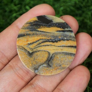 May include: A round, polished, yellow, black, and white banded agate cabochon. The cabochon has a swirling pattern of colors and is approximately 1 inch in diameter.