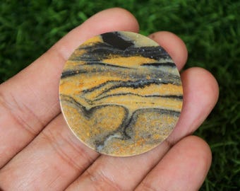 Könnte beinhalten: Ein runder, polierter, gelb-schwarz-weißer gebänderter Achat-Cabochon. Der Cabochon hat ein wirbelndes Muster aus Farben und ist etwa 2,5 cm im Durchmesser.