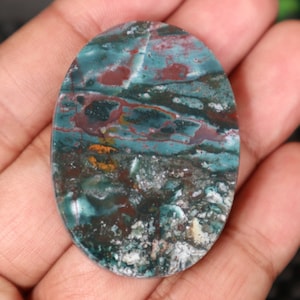 May include: An oval cabochon gemstone with a swirling pattern in shades of green, brown, and white. The stone is polished and has a smooth surface.