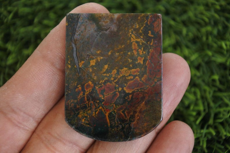 以下が含まれることがあります： 丸みを帯びた下端を持つ長方形の宝石。石は、濃い緑、茶色、赤、黄色などの色の混合物と大理石模様が特徴です。石は手で持たれています。