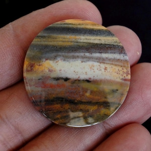 May include: A circular, polished stone displaying a layered pattern of colours. The stone has bands of brown, black, yellow, orange, and white. The stone is held in a hand against a dark background.