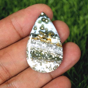 Puede incluir: Un cabujón de jaspe oceánico en forma de lágrima con un patrón blanco, verde y marrón. La piedra tiene una superficie lisa y pulida.