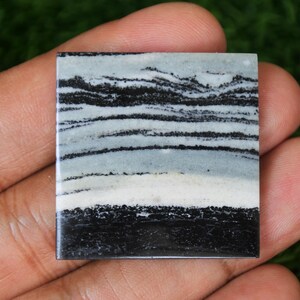 Pode incluir: Um cabochão de ágata quadrado, com faixas pretas e brancas, com uma faixa branca no centro. A pedra tem uma superfície polida e lisa.