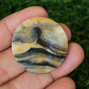 May include: A round, polished stone cabochon with a swirling pattern of black, white, and yellow. The stone has a natural, organic look.