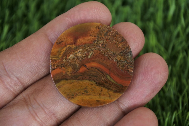 Pu&ograve; includere: Un cabochon rotondo in pietra lucidata con un motivo a spirale di colori marrone, arancio e rosso. La pietra ha un aspetto naturale e organico.