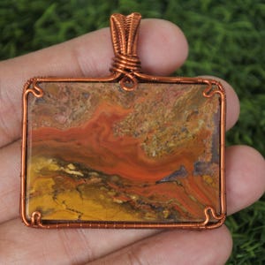 Puede incluir: Colgante rectangular con marco y fianza de alambre de cobre. El colgante presenta una piedra con un patrón arremolinado de tonos naranja, amarillo y marrón. El alambre está intrincadamente envuelto alrededor de la piedra.