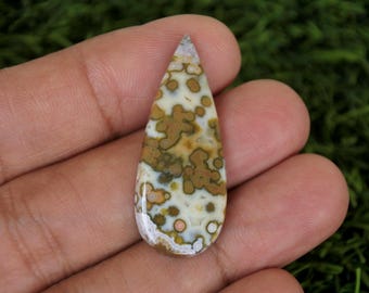 May include: A polished, teardrop-shaped gemstone. The stone has a cream base with brown and tan mottled patterns. The top is light grey. The gemstone is held in a hand against a green background. The stone's length is approximately 5 cm.
