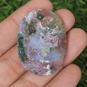May include: Oval shaped cabochon of moss agate with a mix of green, pink, white, and brown colours. The stone has a polished finish and is a natural gemstone.