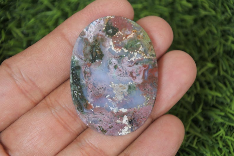 May include: Oval shaped cabochon of moss agate with a mix of green, pink, white, and brown colors. The stone has a polished finish and is a natural gemstone.