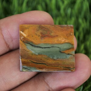 May include: A square, polished, multi-coloured stone with brown, orange, green, and black swirls. The stone is being held in a hand.