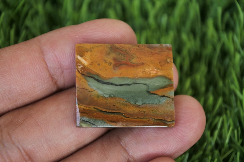 May include: A square, polished, multi-coloured stone with brown, orange, green, and black swirls. The stone is being held in a hand.