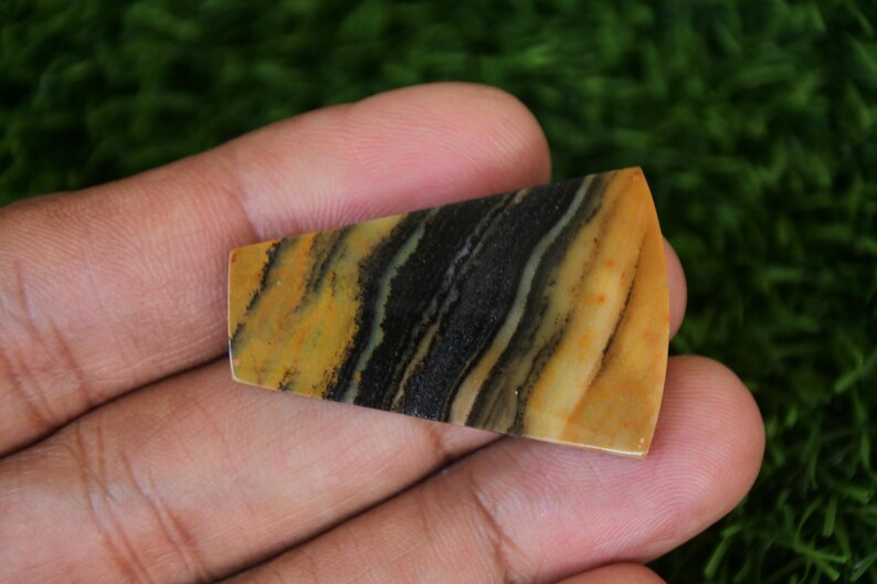 May include: A yellow and black banded agate cabochon with a triangular shape. The stone has a polished surface and is ready to be set in jewelry.