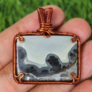Puede incluir: Un colgante rectangular con una piedra gris claro y negra, encerrada en alambre de color cobre. El alambre está intrincadamente envuelto alrededor de la piedra y forma un lazo en la parte superior para una cadena. La piedra tiene un patrón veteado único.