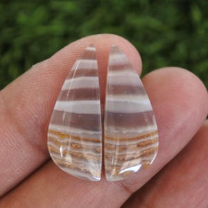 Pode incluir: Duas pedras preciosas em forma de gota com um padrão em camadas de branco, castanho e dourado. As pedras são seguradas entre os dedos, mostrando suas superfícies polidas e variações de cores naturais. Ideais para fazer joias.