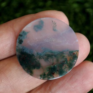 Könnte beinhalten: Ein runder, polierter Edelstein-Cabochon mit einer durchscheinenden, blassrosa Basis, die komplizierte, dunkelgrüne, moosartige Einschlüsse und kleine rotbraune Mineralablagerungen auf der glatten, flachen Oberfläche aufweist.