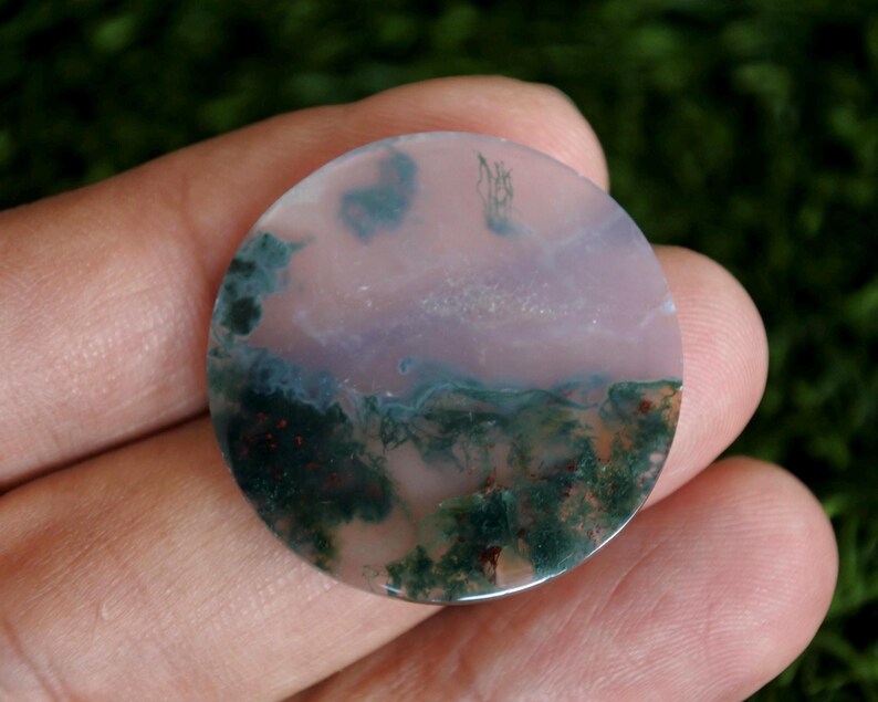 Op de afbeelding: Een ronde, gepolijste edelsteen cabochon met een doorschijnende, lichtroze basis met ingewikkelde, donkergroene mosachtige insluitingen en kleine roodbruine mineraalafzettingen verspreid over het gladde, platte oppervlak van de steen.