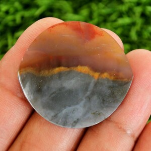 Peut inclure: Une pierre précieuse polie et ronde avec un aspect en couches. La partie supérieure est un mélange de teintes orange et rouge, passant à une bande marron, suivie d'une fine bande jaune, et se terminant par une base grise et blanche. La pierre est tenue dans une main.
