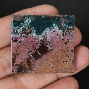 May include: A square, polished, multi-colored stone with shades of green, pink, brown, and white. The stone has a mottled, marbled appearance.