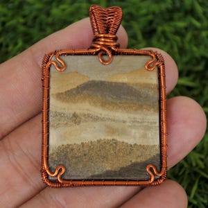 May include: A square pendant with a copper wire frame and a beige and brown agate stone. The stone has a layered pattern that resembles a landscape.