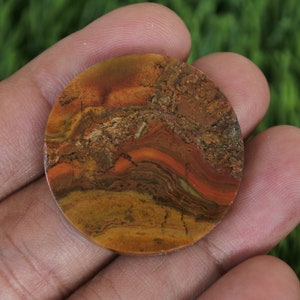 Może przedstawiać: Okrągły, polerowany kamień kaboszonowy z wirującym wzorem w kolorze brązowym, pomarańczowym i czerwonym. Kamień ma naturalny, organiczny wygląd.