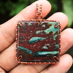 Puede incluir: Un colgante envuelto en alambre de cobre con una piedra cuadrada roja, verde y marrón con un patrón de remolino.