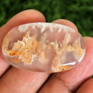 May include: A polished, oval gemstone with a translucent, pale pink base. The stone features intricate patterns of white and golden-orange inclusions, creating a unique, natural design. The gemstone is held in a hand against a blurred green background.