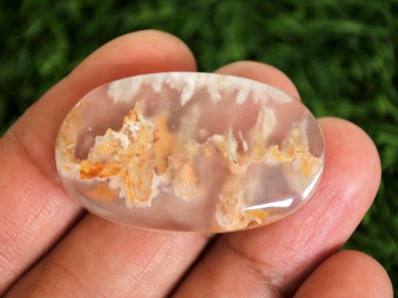 May include: A polished, oval gemstone with a translucent, pale pink base. The stone features intricate patterns of white and golden-orange inclusions, creating a unique, natural design. The gemstone is held in a hand against a blurred green background.