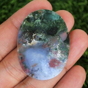May include: Oval-shaped moss agate gemstone with a unique pattern. The stone features a blend of green, white, blue, and red hues. The agate is held in a hand against a blurred green background.