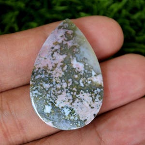 Puede incluir: Un cabujón de ágata rosa y verde en forma de lágrima con un patrón moteado. La piedra está pulida y tiene una superficie lisa.