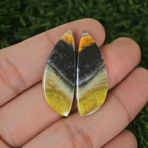 Może przedstawiać: Para caboszonów w kształcie łzy z jaspisu bumblebee w kolorze żółtym, czarnym i białym. Kamienie są wypolerowane i mają gładkie, błyszczące wykończenie.