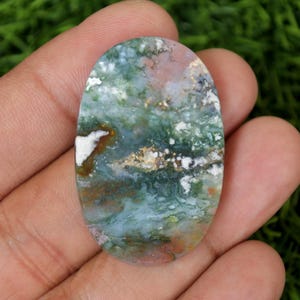 May include: An oval gemstone with a distinctive pattern of green, blue, white, and brown colours. The polished surface shows a landscape-like design, with areas of white and gold. The stone is held in a hand against a green background.