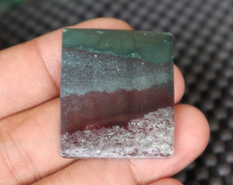 May include: A green, grey, and red banded agate cabochon with a white, cloudy pattern. The cabochon is rectangular and has a smooth, polished surface.