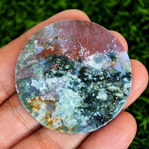 May include: A circular gemstone with a polished surface, displaying a mix of colours. The top section is a reddish-brown, blending into green and blue tones, with white spots. The stone is held in a hand.