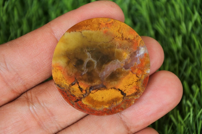 Op de afbeelding: Een ronde, gepolijste agaat cabochon met een mix van gele, oranje, rode en bruine kleuren. De steen heeft een uniek patroon van swirls en aderen.