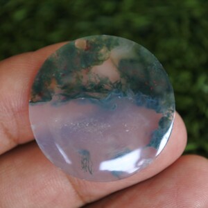 Op de afbeelding: Een ronde, gepolijste mosagaat edelsteen cabochon met een doorschijnende, licht lavendelkleurige basis en opvallende, donkergroene, mosachtige dendritische insluitingen die zich voornamelijk in het bovenste gedeelte van de steen bevinden.