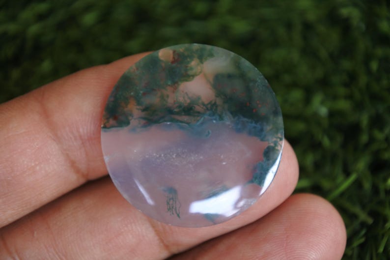 Op de afbeelding: Een ronde, gepolijste mosagaat edelsteen cabochon met een doorschijnende, licht lavendelkleurige basis en opvallende, donkergroene, mosachtige dendritische insluitingen die zich voornamelijk in het bovenste gedeelte van de steen bevinden.