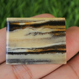 Pode incluir: Um cabochão de ágata quadrada com um padrão em camadas de listras pretas, marrons e brancas. A pedra tem uma superfície polida e lisa.