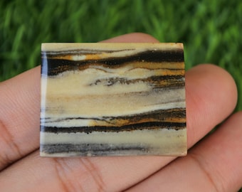 Könnte beinhalten: Ein quadratischer, gebänderter Achat-Cabochon mit einem geschichteten Muster aus schwarzen, braunen und weißen Streifen. Der Stein hat eine polierte, glatte Oberfläche.