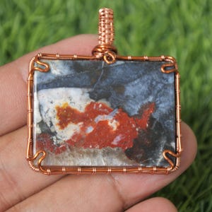 Puede incluir: Colgante rectangular con marco de alambre de cobre. La piedra presenta una mezcla de colores, incluyendo rojo, blanco y azul, con un lazo de alambre de cobre para colgar. El colgante se sostiene en una mano sobre un fondo verde.