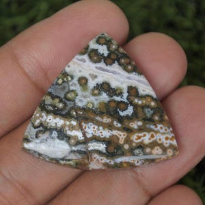 Puede incluir: Una gema triangular con un patrón único de manchas y remolinos marrones, blancos y beige. La piedra pulida se sostiene en una mano, mostrando sus colores y texturas naturales. El diseño de la piedra recuerda a un paisaje.
