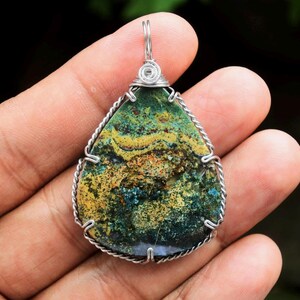 May include: A teardrop-shaped pendant with a silver wire frame. The stone features a mix of green, yellow, and brown hues. The pendant is held by a silver wire loop for a necklace.