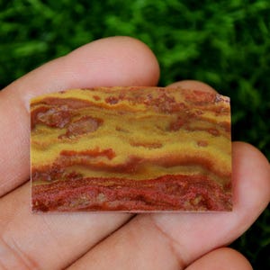 Pode incluir: Um close-up de um cabochão de ágata vermelho e amarelo. A pedra tem um padrão em camadas, estriado, com uma superfície lisa e polida.