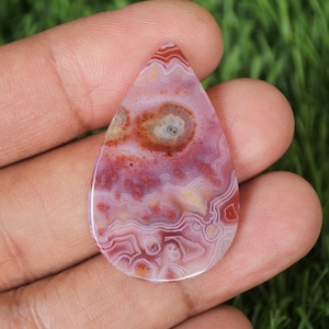 Puede incluir: Un cabujón de ágata en forma de lágrima con un patrón de color rosa, blanco y marrón. La piedra tiene un diseño de espiral y capas con un ojo central.