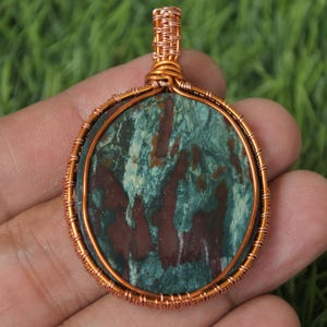 Może przedstawiać: Zawieszka owinięta miedzianym drutem z zielonym i brązowym agatem o wirującym wzorze.