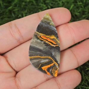 Könnte beinhalten: Ein tropfenförmiger Cabochon aus poliertem Pyrit mit einem schwarzen, goldenen und orangefarbenen Muster. Der Stein ist etwa 5 cm lang.